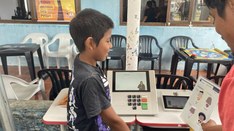 Uma criança sorridente está em frente a uma urna eletrônica, simulando um voto, enquanto um adul...