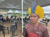 Um homem indígena com cocar de penas amarelas e azuis, vestindo uma camiseta marrom, olha direta...