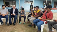 Um grupo de pessoas está sentado em cadeiras de plástico ao ar livre, em frente a um prédio de c...