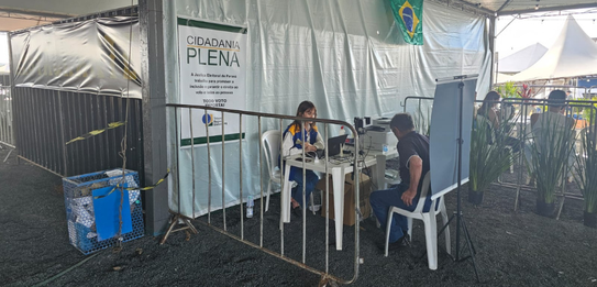 Fotografia que retrata o atendimento eleitoral em uma sala de aula adaptada. Dois cidadãos são a...
