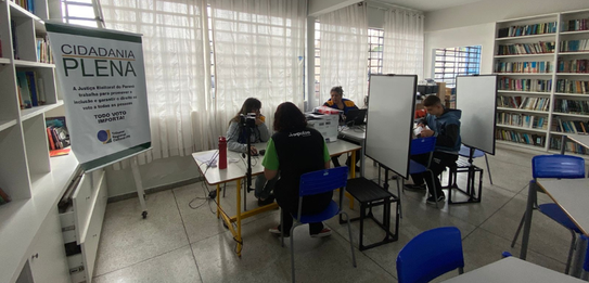 Fotografia de um homem e duas mulheres sentados em cadeiras azuis diante de mesas brancas sobre ...