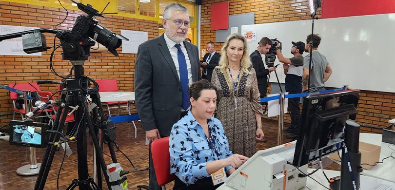 Fotografia de um ambiente de auditoria, em uma sala de aula. Há uma mulher em frente a uma urna ...