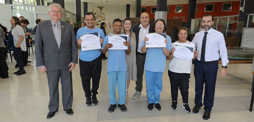Fotografia em que são vistos dois homens e duas mulheres segurando um diploma, posando ao lado d...