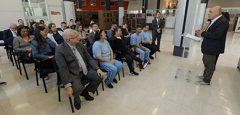 Fotografia em que se vê, do lado direito, um homem discursando para um público sentado em cadeir...