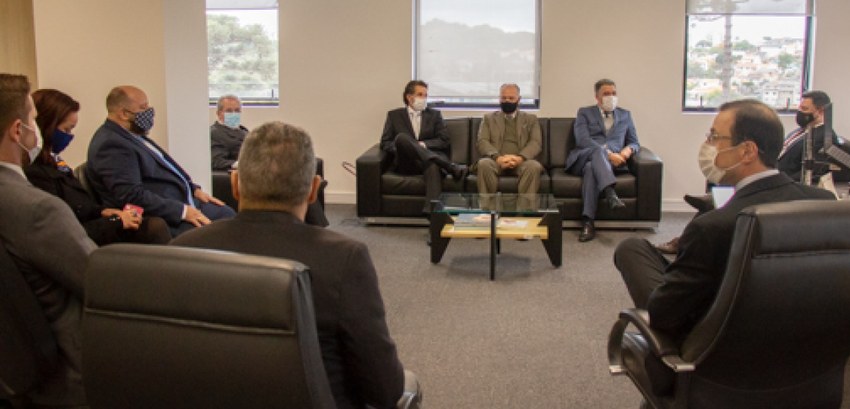 Fotografia de homens de terno e gravata sentadas em sofás e poltronas pretas em uma sala