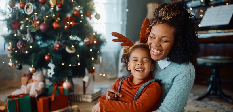 Foto de mãe e filha abraçadas sorrindo. A mãe usa blusa azul e a filha, vermelha, com adereço ve...