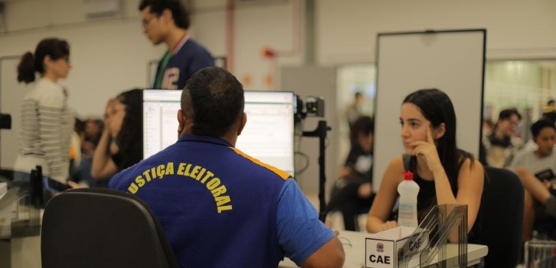 Fotografia em plano médio de um atendimento ao público da Justiça Eleitoral em um galpão amplo. ...