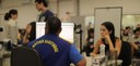Fotografia em plano médio de um atendimento ao público da Justiça Eleitoral em um galpão amplo. ...