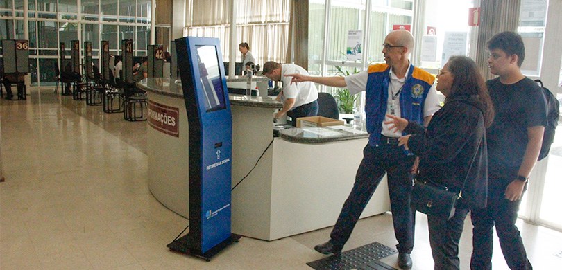 Fotografia que retrata o atendimento em um ambiente institucional. Em primeiro plano, à direita,...