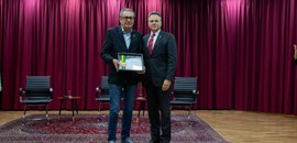 Fotografia em que aparecem dois homens posando no palco do auditório do TRE-PR. Um deles mostra ...