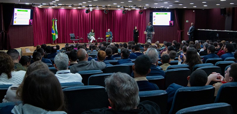 Fotografia em que se observa, ao fundo, o palco do auditório do TRE-PR, sobre o qual veem-se uma...