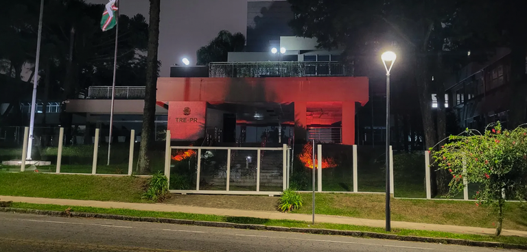 Fotografia da sede do TRE-PR à noite com iluminação na cor vermelha. O prédio está atrás de um m...