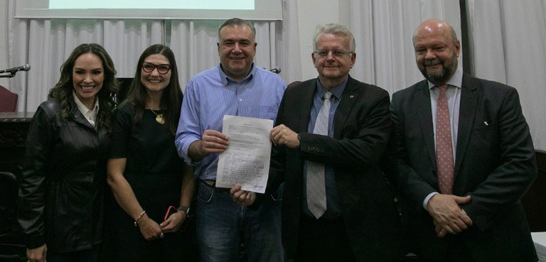 Fotografia de duas mulheres e três homens posando sorrindo. Ao centro, dois homens seguram junto...