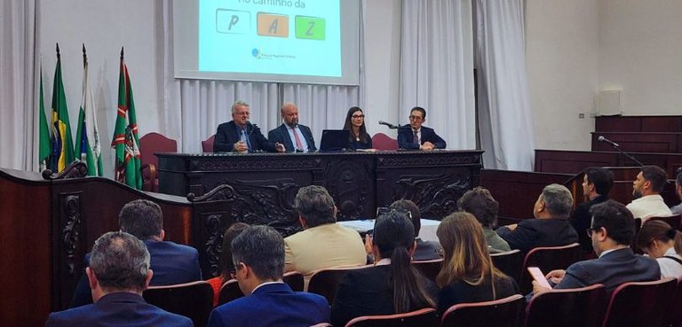 Fotografia da mesa de autoridades, composta por três homens e uma mulher, no palco de um auditór...