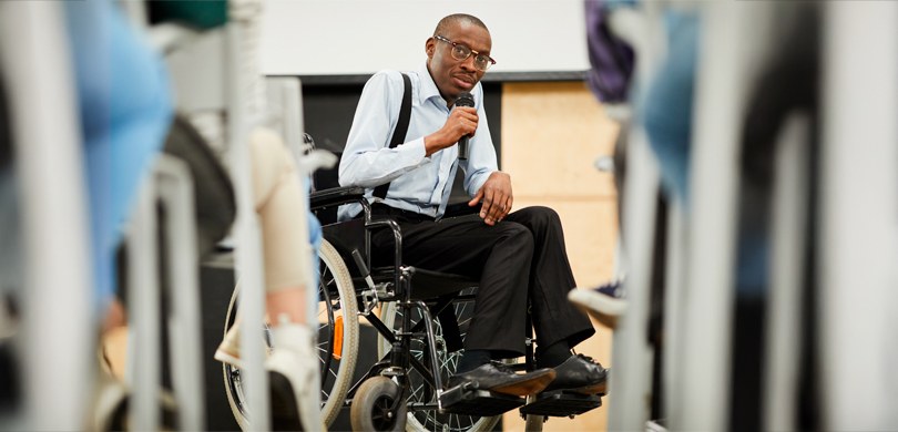 Fotografia de um homem em uma cadeira de rodas segurando um microfone. Ele é uma pessoa negra, q...
