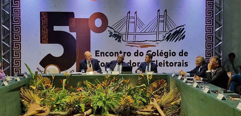 A fotografia é da mesa de autoridades do evento. Ao fundo existe um painel em que está escrito: ...