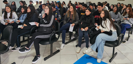 Fotografia em que se observa um grupo de jovens, homens e mulheres, sentados em um auditório ou ...