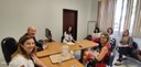 Fotografia de seis mulheres e um homem sentados ao redor de três mesas durante uma reunião. Sobr...