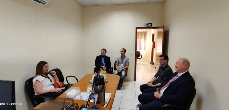 Fotografia de uma mulher e quatro homens ao redor de uma mesa durante uma reunião. Sobre a mesa,...