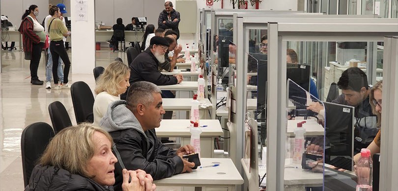 Fotografia que retrata um ambiente interno de atendimento ao público com diversas pessoas sentad...