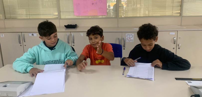 Fotografia de três meninos sentados atrás de uma mesa, atuando como mesários na eleição do Proje...