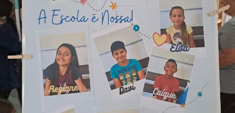 Fotografia de um cartaz branco, em que se lê em azul: “A Escola é Nossa!”. Ao redor, há quatro f...