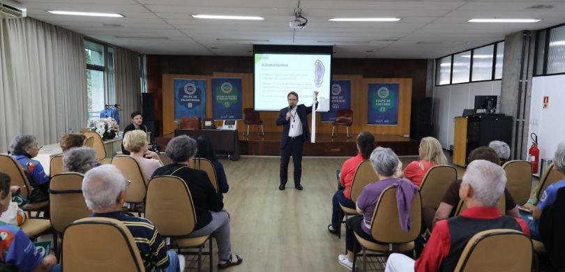 Fotografia em auditório na qual um homem veste terno escuro, camisa clara e crachá ao pescoço, e...