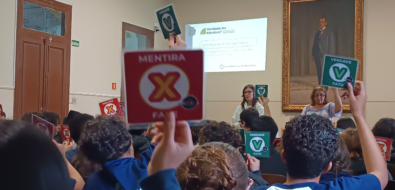 Fotografia em que se observa um auditório com diversas pessoas de costas em relação à câmera, er...