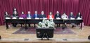 A foto é da mesa de autoridades. São onze pessoas sentadas atrás de uma mesa retangular grande. ...