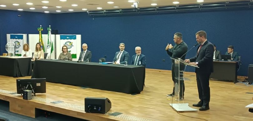 Fotografia de uma solenidade oficial sobre o palco de um auditório com piso de madeira e parede ...