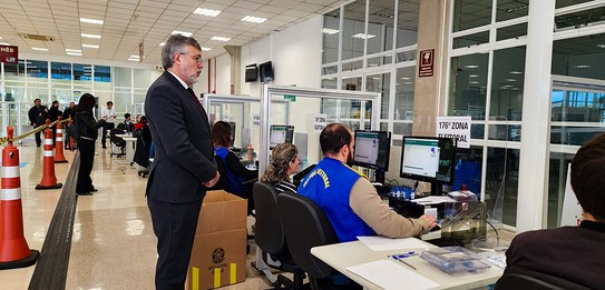 Fotografia de uma série de guichês de atendimento. Há um homem de terno e gravata em pé observan...