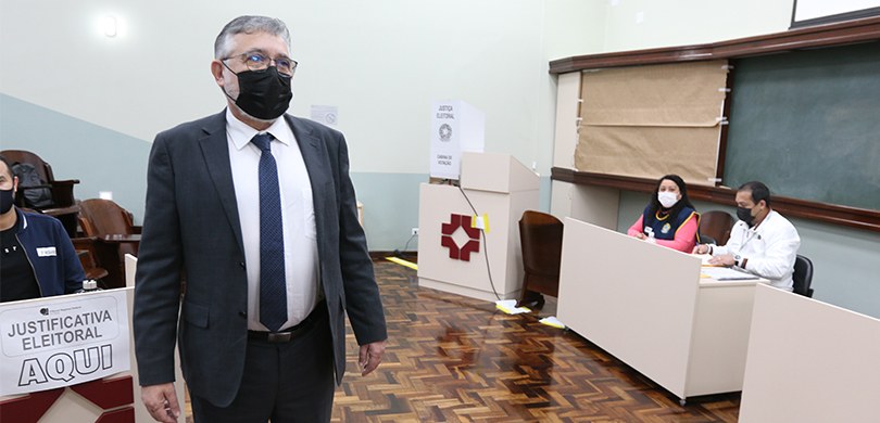 Fotografia de um homem de terno e gravata em uma sala. Há uma cabine de votação ao fundo, ao lad...