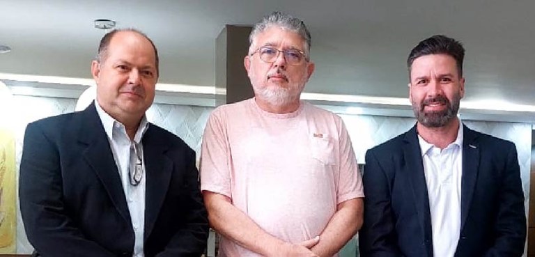 Três homens posam em uma sala, com piso de granito. Os dois que estão nas pontas usam calças jea...