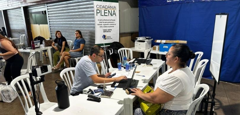 Cidadania Plena atende eleitores na rodoviária de Maringá até este sábado (25) Fotografia em que se vê um homem atendendo uma eleitora. Eles estão sentados em cadeiras brancas...