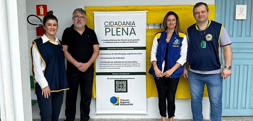 Fotografia de duas mulheres e dois homens posando sorrindo ao lado de um banner em que se lê: Ci...