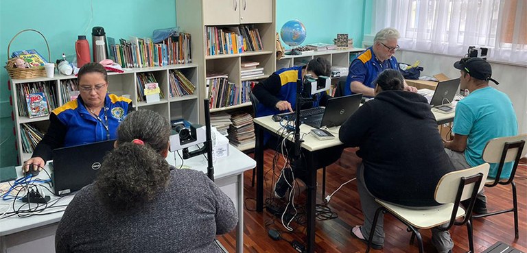 Fotografia de uma mulher e um homem com o colete da Justiça Eleitoral  atendendo duas mulheres e...