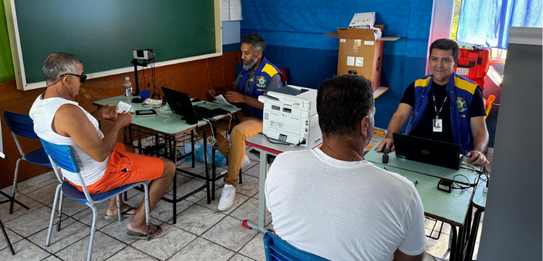Fotografia que retrata o atendimento eleitoral em uma sala de aula adaptada. Dois cidadãos são a...