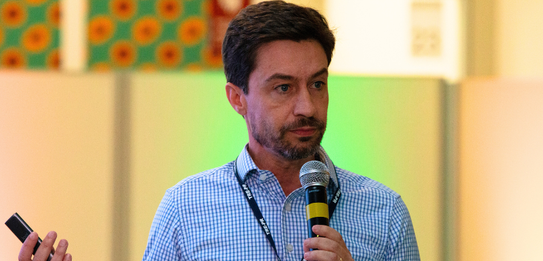 Fotografia de nove pessoas, entre homens e mulheres, posando sorrindo em frente a um edifício co...