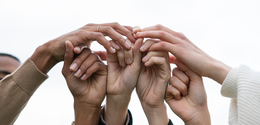 Fotografia que retrata diversas mãos de diferentes tons de pele unidas e entrelaçadas ao centro ...