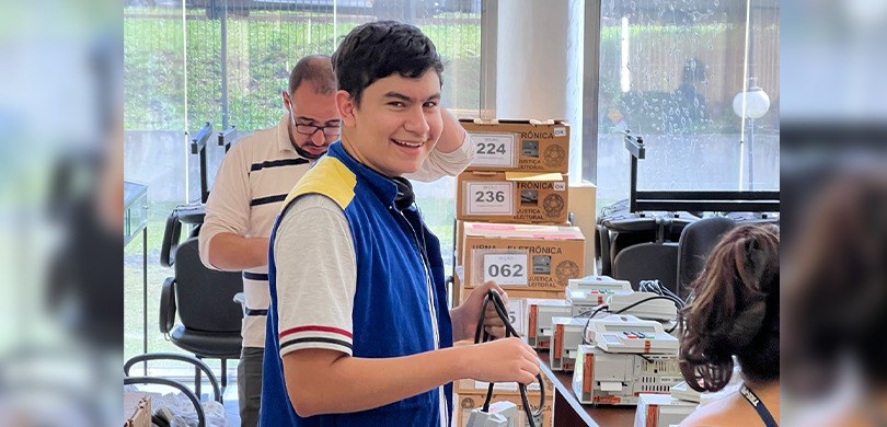 Fotografia de um jovem de cabelos escuros sorrindo para a foto. Ele  veste o colete da Justiça E...