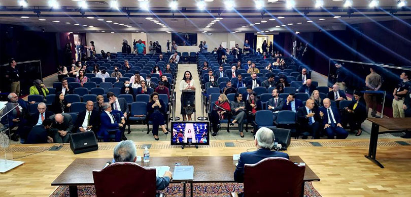 Fotografia de um auditório com assentos em tom de azul. Dois homens estão sentados à mesa em cim...