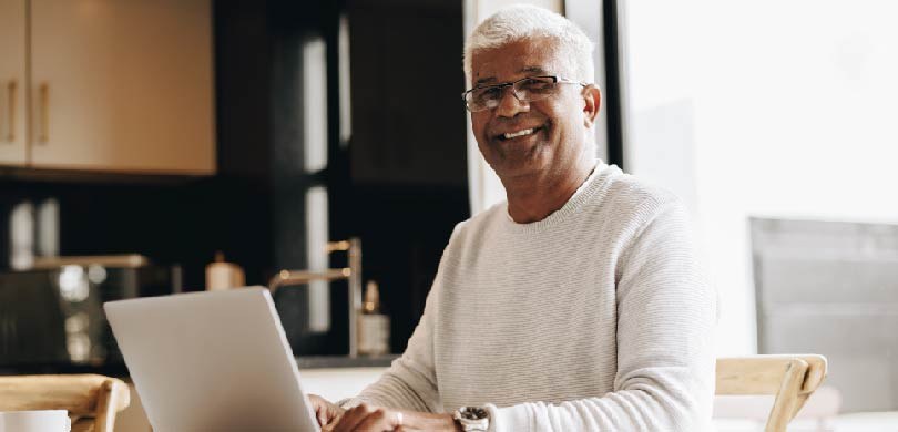 Um senhor idoso mexe no computador, que está em cima da mesa. O homem de pele negra e cabelos br...
