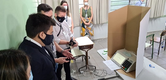 Começa a auditoria de funcionamento da urna eletrônica Foto de uma cabina de votação com urna eletrônica em cima de uma mesa dentro de uma sala de aula...