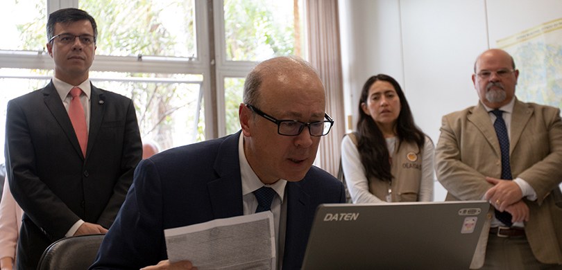 Fotografia de um senhor de terno e gravata e óculos de leitura em frente a um notebook. Ao seu r...
