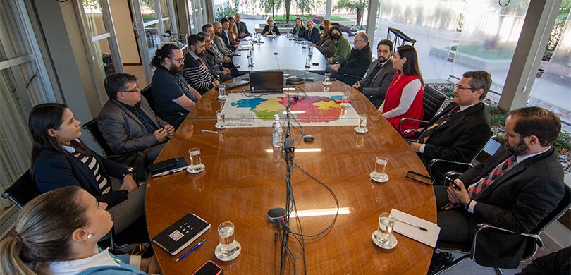Fotografia de um grupo de diversas pessoas reunidas ao redor de uma mesa, durante reunião.