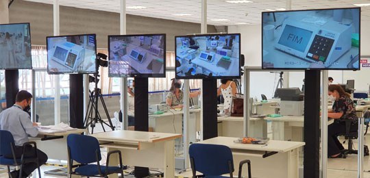 Fotografia de um ambiente de auditoria. Há cinco televisões em primeiro plano com imagens de urn...