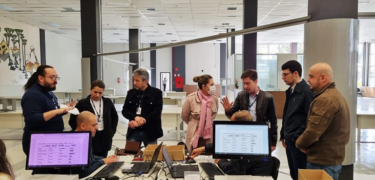 Fotografia de um grupo de pessoas, entre homens e mulheres, ao redor de uma mesa com notebooks e...