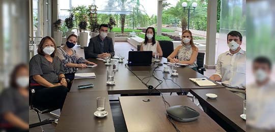Fotografia de um grupo de pessoas em volta de uma mesa de reuniões. Todas usam máscara e olham p...