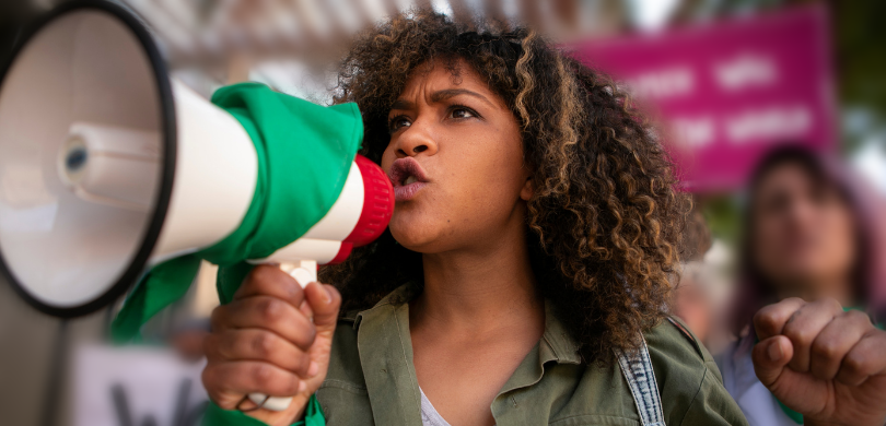 Fotografia que retrata um protesto em ambiente externo com foco em uma mulher jovem de pele negr...