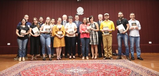 Fotografia de um grupo de pessoas em cima de um palco. Todas seguram um livro. Atrás delas há um...